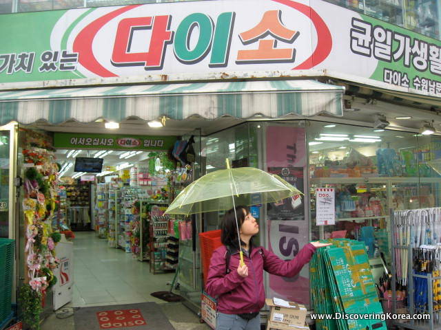 A woman carrying a green umbrella walks out of a shop, in the background the inside of the shop is visible, and to the top of the frame Korean lettering.