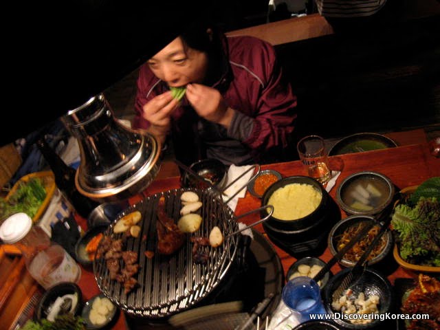 A woman sitting at a table filled with food in various bowls, eating. In the center of the frame is a tabletop grill containing meat.