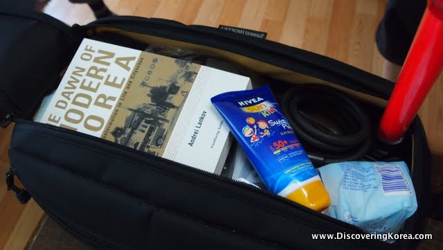 A black cycling saddle bag, packed with a book, sunscreen and and other items below. On a wooden floor.