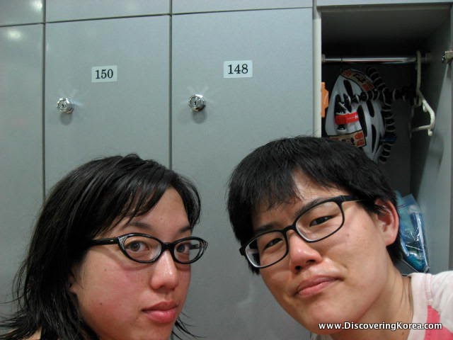 Close up of two women's faces in front of an open locker on the right and closed lockers on the left of the frame.