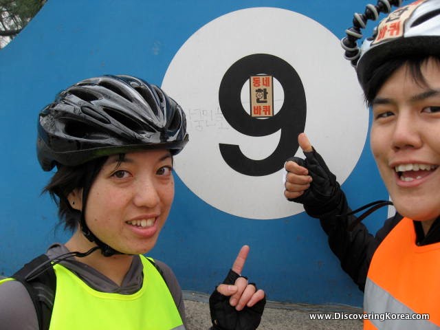 Two women pictured wearing bicycle helmets and reflective safety vests pointing at a number 9 on a blue and white wall.