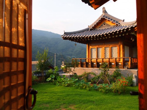 An ornate Korean house in the sunshine with mountain backdrop, green lawn to the front.