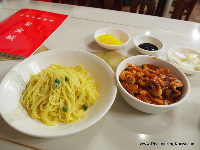 Jjajangmyeon noodles pictured in a white bowl, with a seafood sauce, and condiments in white bowls on a white surface. A red menu is to the left of the frame.