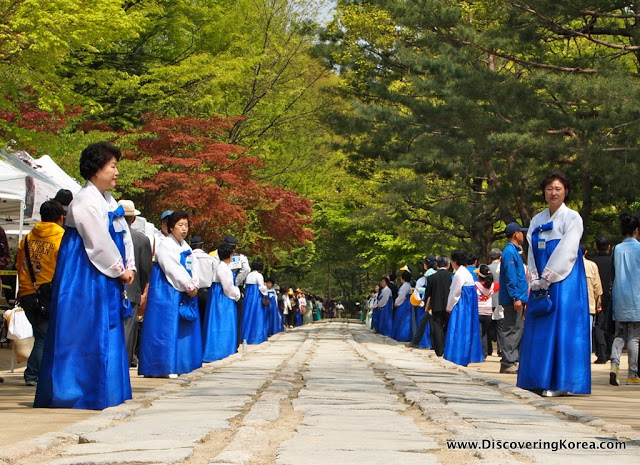 Two lines of women, standing opposite each other, dressed in white and blue robes with trees in the background and a stone path in between, for the Jongmyo Great Rite.