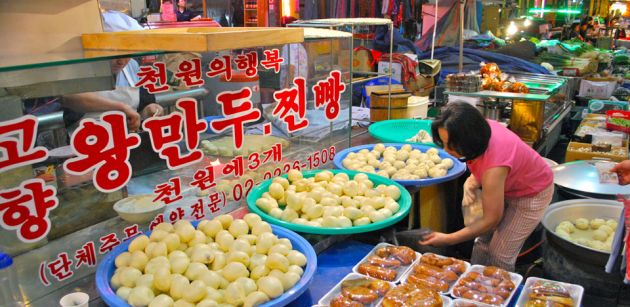 A food vendor's stall at Jungang Traditional Market in Seoul, various vegetables and meat produce in plastic bowls, with a glass cabinet and red Korean writing. A woman in the foreground inspects the wares.