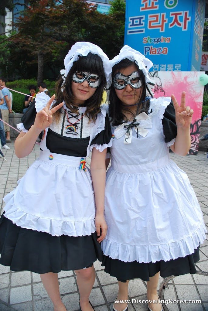 Two Korean women, dressed in black dresses with white aprons, and face masks pose in front of a blue sign.