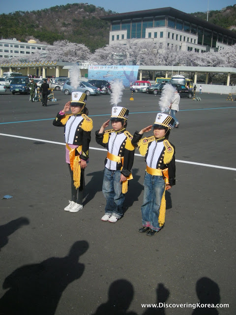 Three Korean middle school students in marching band uniforms are saluting.