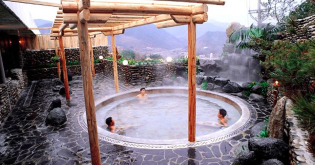 An outdoor hot spa pool, under a wooden pergola, set into tiled patio with soft focus mountains in the background.