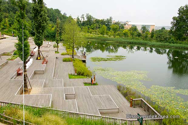 Geometric steps with bench seating, ornamental trees, leading down to the lake at West Seoul Lake Park. The clear water of the lake reflects the trees and grasses in the background that surround it.