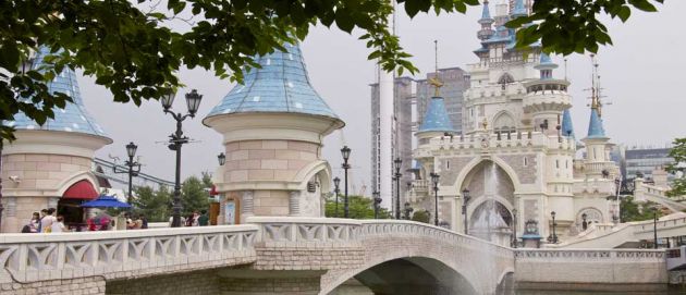 View across a bridge to the impressive stone building of Lotte World Amusement Park. Circular turrets with light blue roofs and an arched entranceway, it looks like a castle.