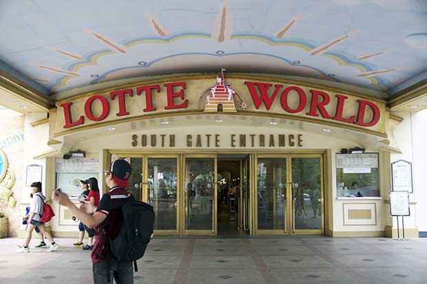 Entrance to Lotte World, glass doors surrounded by a light yellow wall with red lettering, in front of the entrance various people walking and standing around.