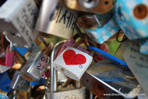A close up of a cluster painted padlocks, with Korean lettering, and to the center of the frame, one painted with a white background and a red love heart.