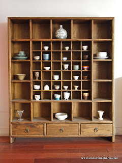 Ceramic rice beer cups and ceramic vases in a wooden display cabinet, with drawers at the bottom, standing on a wooden floor.