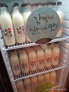 A display refrigerator containing bottles of Makgeolli rice beer. A sign with Korean lettering on the front of the fridge.