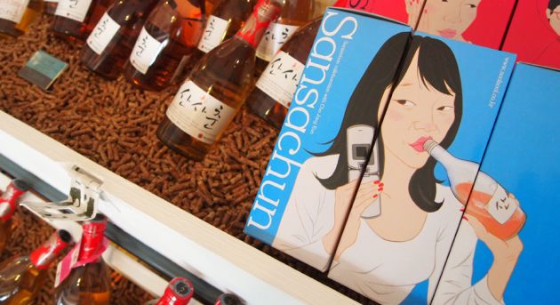 Bottles of rice beer to the left of the frame, with a picture of a woman drinking from a bottle, holding an old fashioned cell phone on a blue background.