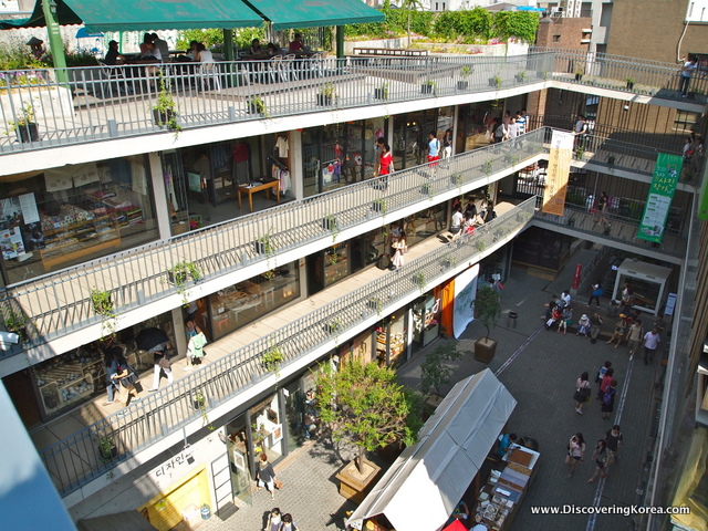 Looking down at a shopping mall, open balconies on three different levels, with shops and cafes, and the road below.