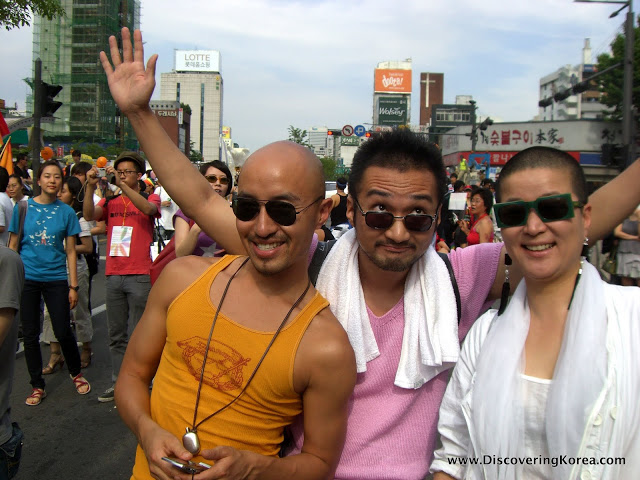 Two men and a woman at a parade, one man with his arms in the air celebrating, with shops and city scene behind.