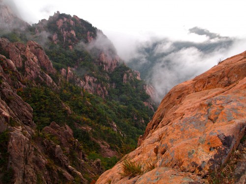 View towards a mountain in Korea with wispy clouds in the background, and forested, rocky slopes.