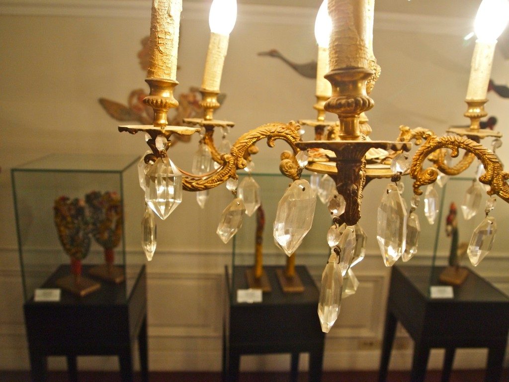 Close up of a chandelier, gold colored with crystals hanging down. In the background are glass display cases at the Musee Shuim.