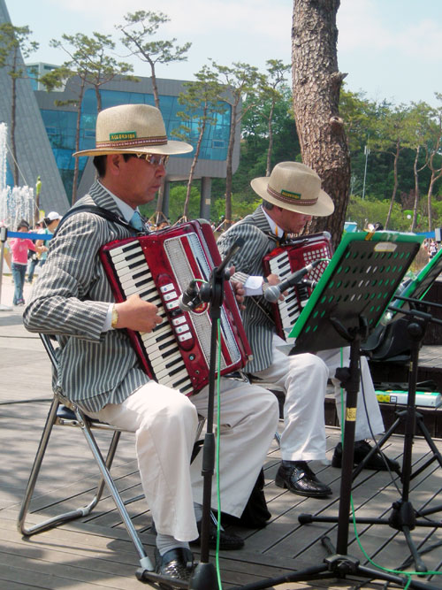 Two musicians, seated, playing keyboard, outdoors in the sunshine at Hampyeong.