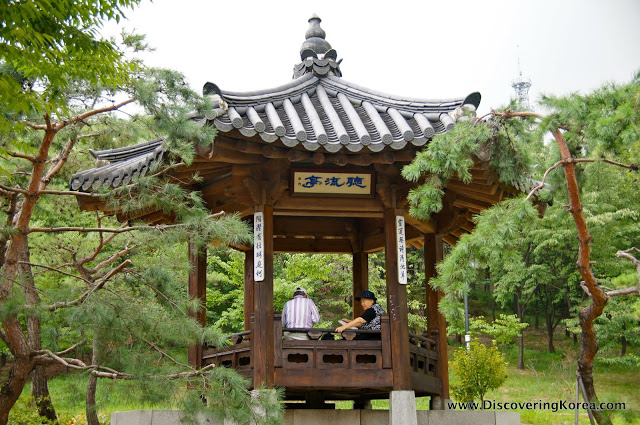 A pergola in Namsan botanical garden. Wooden house with ornate roof and open sides, on a stone plinth surrounded by trees. Two people are sitting in the pergola.