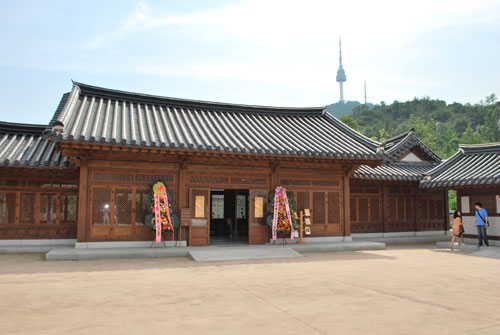 The exterior of Namsangol Hanok village, a brown wooden building with a grey roof, Seoul's tower in the background. Two people to the right, on the stone steps.