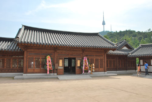 Outside view of Namsangol Hanok, a brown wooden building with open doors and flower decorations outside. Two people to the right of the frame and soft focus tower and vegetation in background.