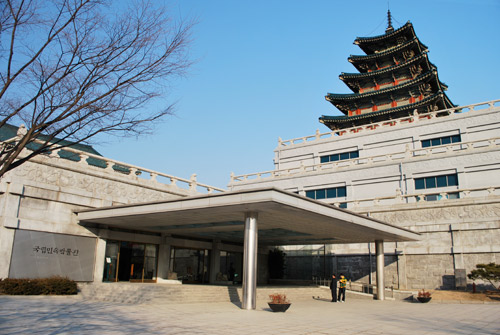 Outside view of the National Folk Museum, a concrete brick building with a low roof over the entrance. A higher, more ornate roof shows behind.