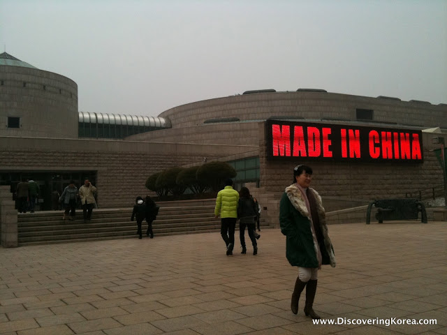 Exterior view of National Museum of Art, a brutalist style stone building with a large red and black 'Made in China' sign to the right. Pedestrians walking towards the steps up to the building.