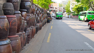 A street in Noksapyeong, with green buses on the right of the frame on a tarmac road, and on the left a long line of large ceramic pots stacked on top of each other.