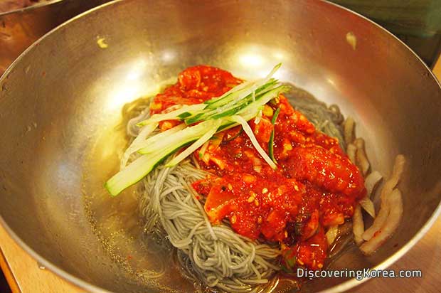 Close up of a metal bowl with noodles, sliced cucumber and a bright red sauce.