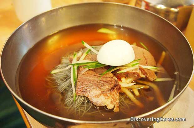 Close up of a metal bowl with a clear broth, containing thin noodles, pieces of meat, with garnish of thinly sliced cucumber and half a boiled egg.