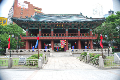 Stone steps leading up to a large building with red pillars and a traditional curved roof, painted in ornate turquoise in the eaves, lawn either side, and flags.