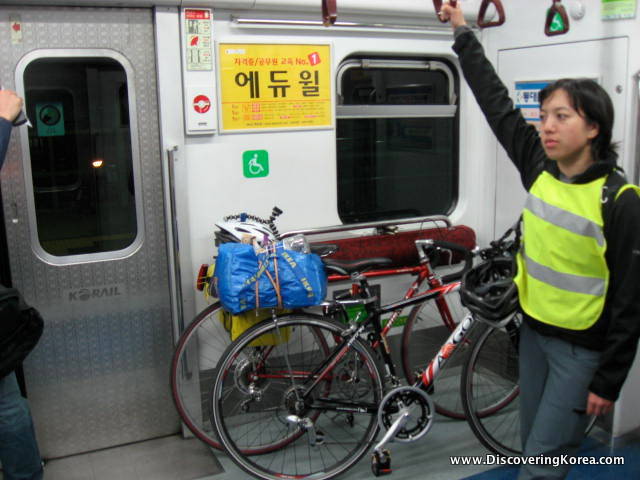 A woman rides the subway with two bicycles, the background is the subway doors and Korean signs.