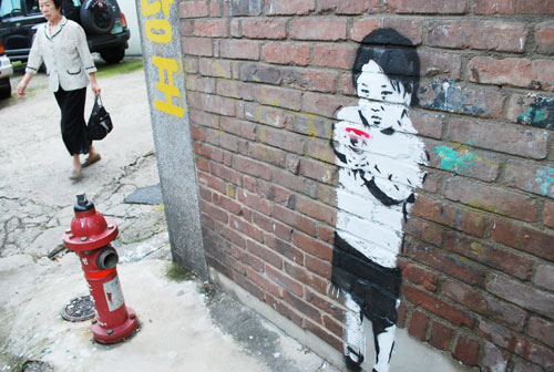 A brick wall with a painting of a child holding a gun, the painting is mostly white, with the child's hair in black. To the left is a red fire hydrant and a pedestrian in the background.