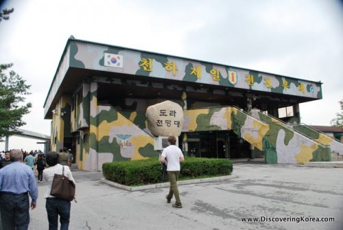 Outside the Dora Observatory in DMZ, the building painted green and yellow camouflage with pedestrians on the left of the frame.