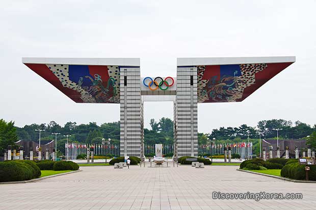 Seoul's Peace Gate at the Olympic Park. A large structure with a flat top, and an entrance in the middle, in front is a paved wide walkway flanked by low bushes and grass.