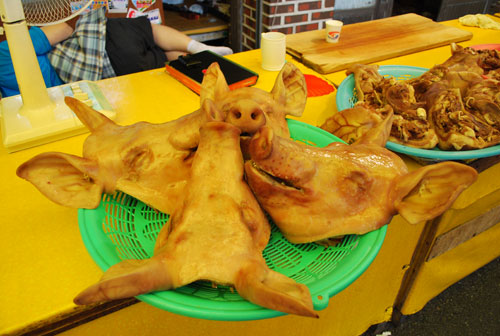 Four pig's heads arranged on a green plastic dish, on a yellow wooden surface, with cooked meat on a blue plastic plate to the right of them.