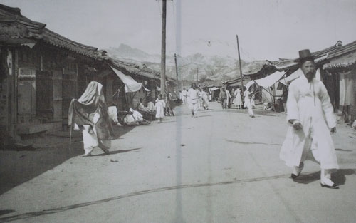 A black and white historical photo of people walking in a street in old Seoul, with traditional houses either side.