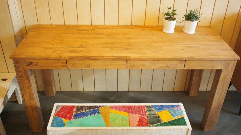 A pine table, in front of a wooden wall, with two small white pots containing plants. In front is a bench seat with multicolored pattern on a white frame.