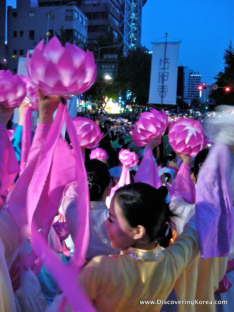 Close up of a woman in yellow, dancing amongst a crowd, with pink, lit-up lotus flowers on an evening backdrop of Seoul's buildings.