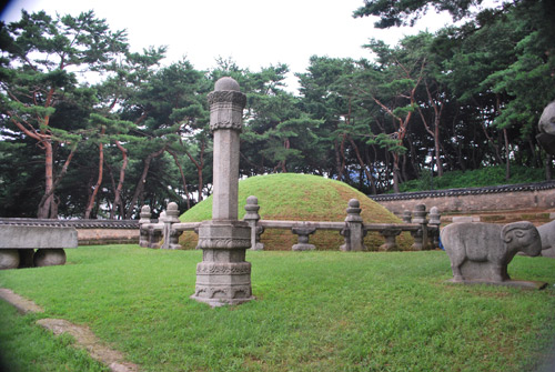 A circular mound covered in grass, surrounded by stone pillars and stone carvings of animals housing Queen Jeonghyeon's tomb. In the background is a pine forest.