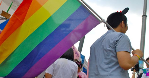 A man in a light blue t shirt carrying a rainbow flag over his shoulder, walking through a crowd of people.