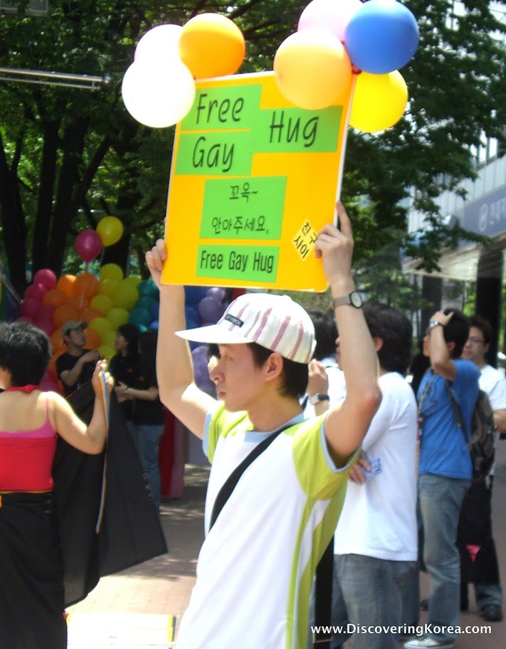 A young man with a white and green t shirt holds up a yellow banner with black writing on a green background and balloons, amongst a crowd of people at Queer Culture Seoul.