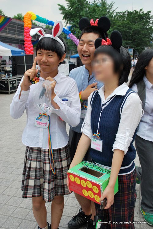 Three young Korean girls dressed in white shirts and plaid skirts, all wearing bunny ears and holding a red and green box.