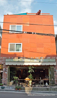 An orange building with two windows and an open storefront, showing table and chairs in sunshine.