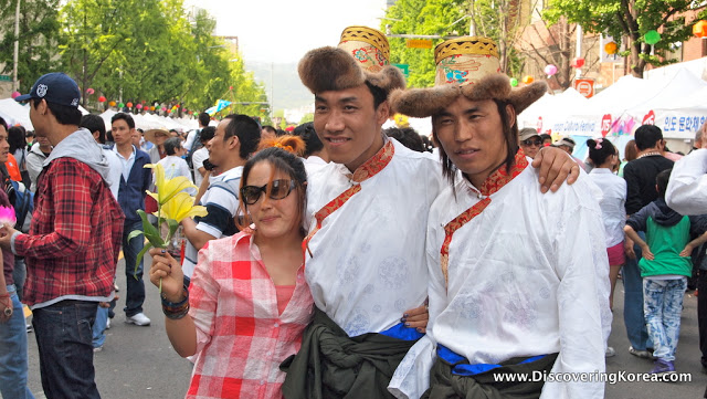 Two men wearing white shirts and yellow decorated hats, with a woman in a checked shirt, pose for the camera in front of street crowds at the Lotus Lantern Festival in Seoul.