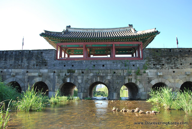 A stream runs under arches below the walls at Suwon fortress. An ornate roof with red and blue coloring at the top of the frame, and river vegetation to the left and right.