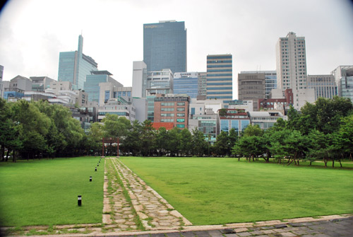 View over a stone pathway with grass and trees either side, towards the city buildings in Seoul.