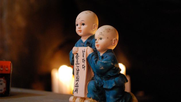 Two dolls, both dressed in blue outfits, one standing, one kneeling, holding up a sign with Korean lettering, on a dark, soft focus background.